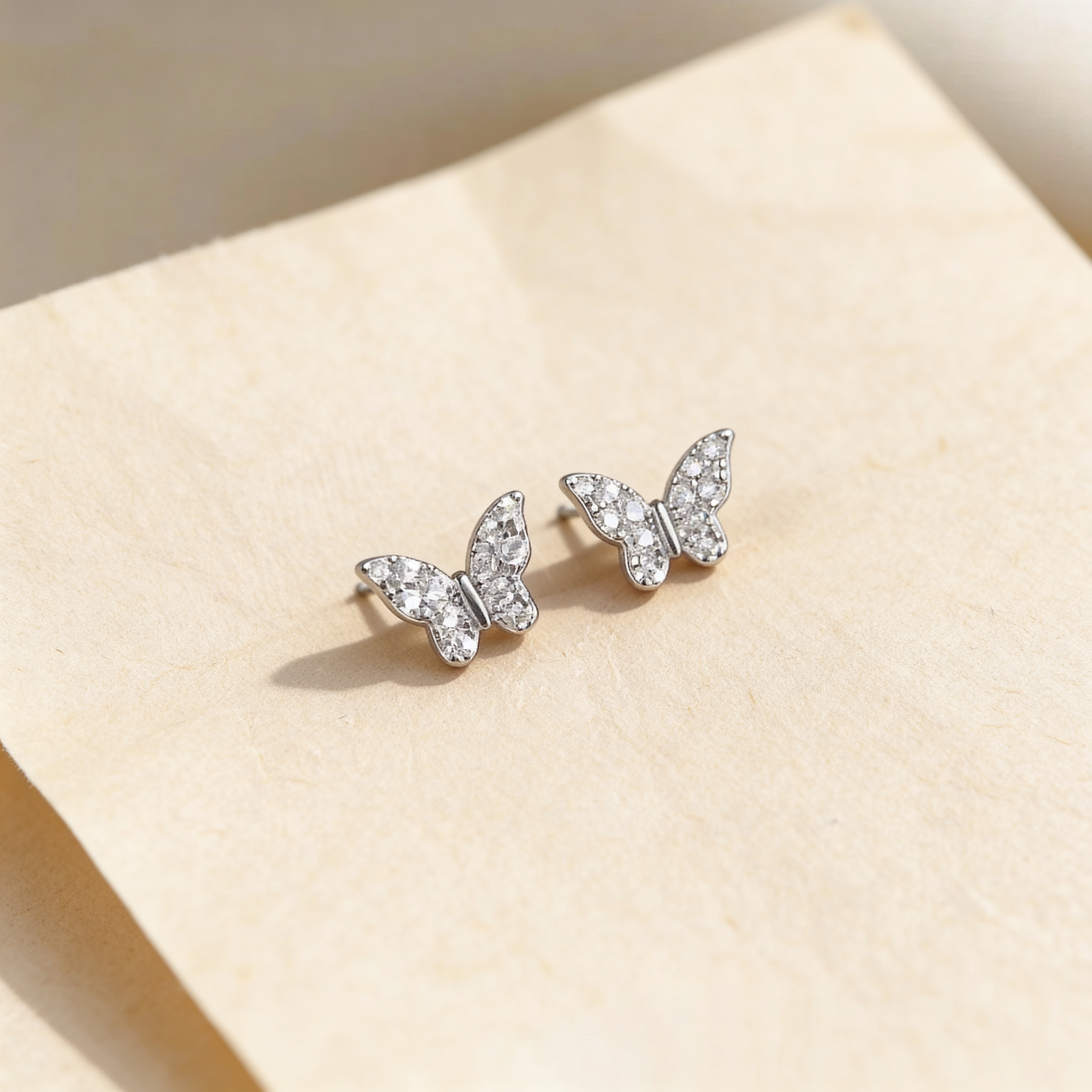 Mini silver plated butterfly stud earrings with tiny sparkling crystal stones on a soft beige background