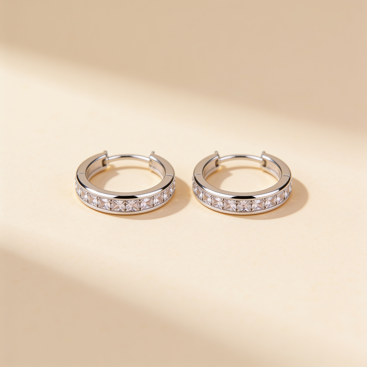 Pair of silver plated crystal huggie hoop earrings with sparkling stones displayed on a warm beige background