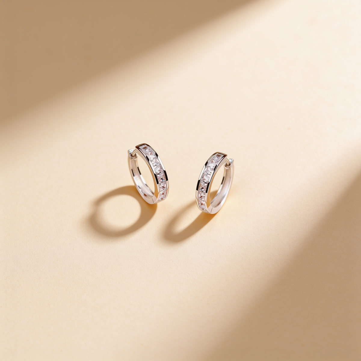 Silver plated crystal huggie hoop earrings with sparkling stones displayed upright on a warm beige background with soft shadows