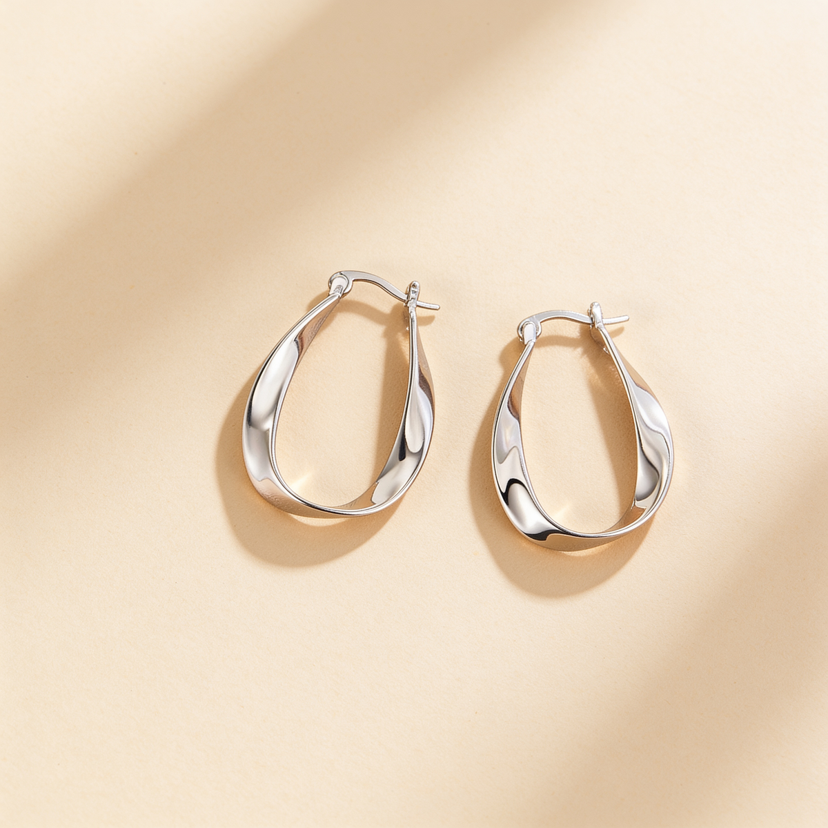 Pair of silver plated twisted hoop earrings with a polished sculpted design displayed on a warm beige background